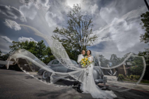 Houston bride and groom pose in front of a vintage car as the bride’s long veil flows dramatically in the wind, creating an elegant and timeless wedding portrait.